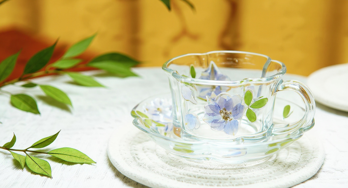 Handmade glass cup and saucer with pressed flowers beside tea and botanical journal