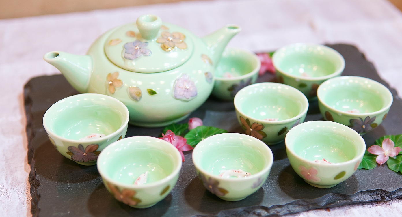 Handmade celadon tea set with teapot and cups on wooden table with jasmine sprigs and linen cloth.