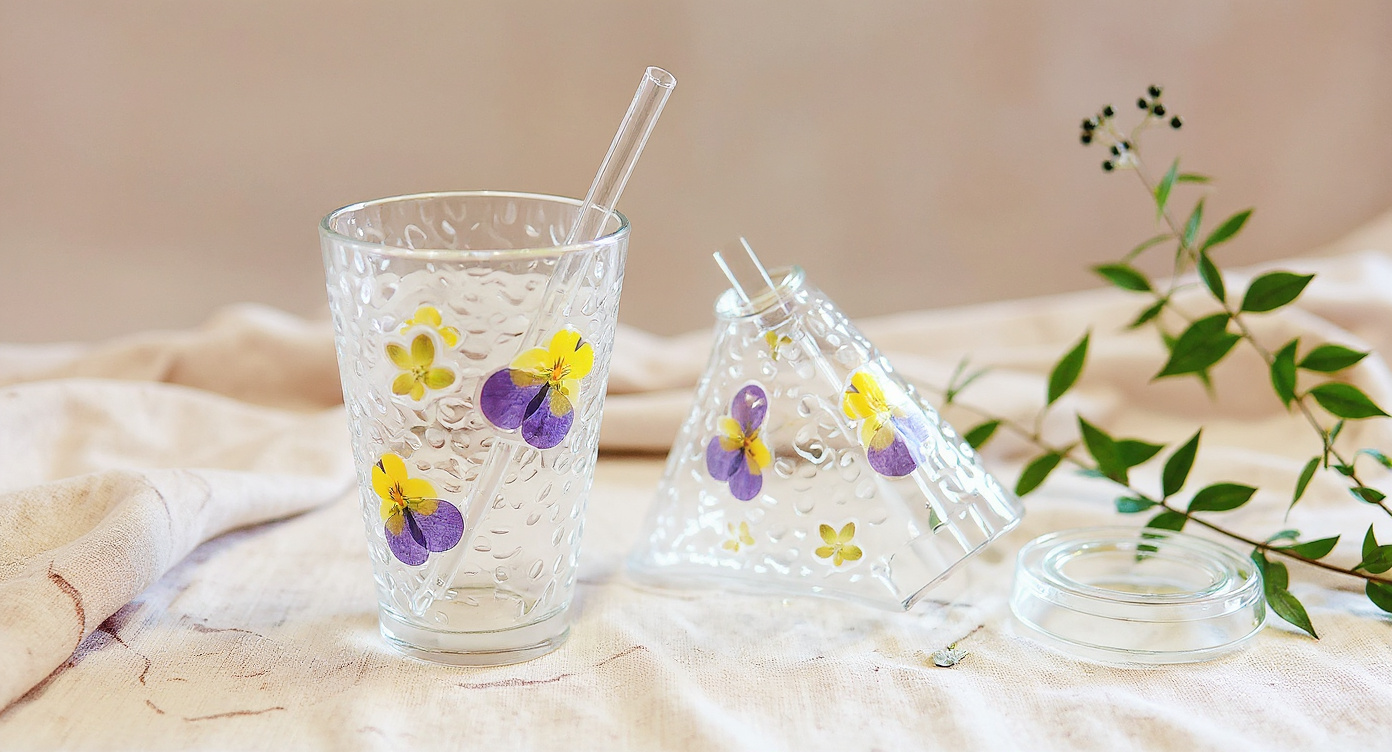 Handmade Floral Glass Cup with warm tea, spoon on saucer, eucalyptus background.